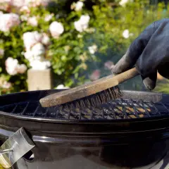 Main tenant gantée, brosse métallique pour nettoyer un barbecue. Des roses roses en arrière-plan.