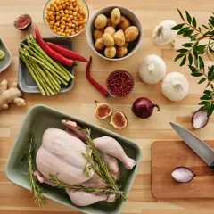 A whole raw chicken in a green dish surrounded by fresh vegetables and herbs on a wooden table.