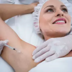 Kobieta z uśmiechem poddaje się zabiegowi leczenia nadpotliwości pach. Lekarz wykonuje zastrzyk.