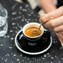 Ręka miesza łyżeczką aromatyczne espresso w czarnej filiżance. Obok szklanka wody. Prosty przepis na espresso jak zrobić.