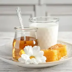 Un bicchiere di latte, un vasetto di miele dorato con un cucchiaino, e favi di miele su un piatto con un fiore bianco. Alcuni credono che latte caldo e miele faccia male.