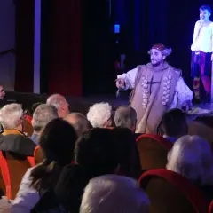 Attori sul palco, uno con un costume particolare, interagiscono con il pubblico, rompendo la quarta parete.