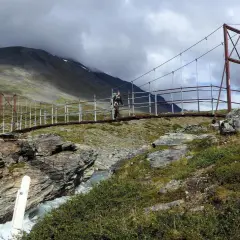 Hängebrucke über einem Fluss im schwedischen Gebirge. Ein Wanderer überquert die Brücke, umgeben von felsiger Landschaft und grüner Vegetation.