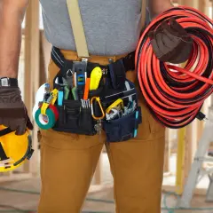 Elektryk z pasem narzędziowym, w którym znajdują się podstawowe narzędzia elektryka: kombinerki, taśma izolacyjna, klucze. W ręku trzyma żółty kask i zwinięty kabel.