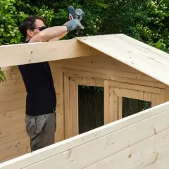 Un homme assemble une cabane en bois, utilisant une perceuse pour fixer une planche sur le toit. La peinture sur bois est visible sur les murs et le toit.