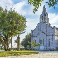 La iglesia de la Toja, cubierta de conchas, se alza majestuosa bajo un cielo azul con nubes. Un árbol frondoso y palmeras la rodean.