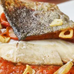 Bacalao a la riojana con piel crujiente, pimientos rojos y ajo frito, servido sobre una cama de salsa de tomate.
