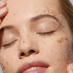 Una cara llena de granos de exfoliante, con dedos aplicándolo suavemente.