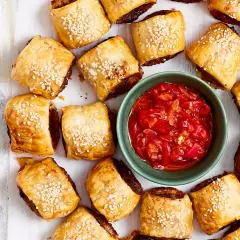 Goldbraune Würstchen im Blätterteig mit Sesam, daneben eine Schale mit roter Chutney-Sauce. Perfekt für schnelle Partysnacks zum Vorbereiten!
