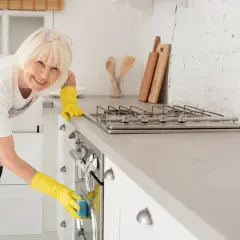 Uśmiechnięta starsza pani w żółtych rękawiczkach czyści kuchnię, pokazując, czym odtłuścić płytki.