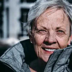 En kvinna med korta frisyrer, dam 65, ler varmt. Hennes grå hår ramar in ett ansikte med vänliga rynkor och blå ögon.