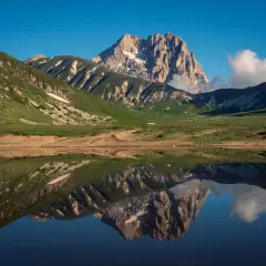 Spiegelglatte See reflektiert majestätisches **Gebirge in Italien** unter blauem Himmel. Grüne Hänge und felsige Gipfel.