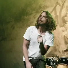 Chris Cornell z zespołem na scenie, w białej koszulce, śpiewa do mikrofonu. W tle perkusja i światła.