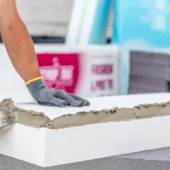 Ręce w rękawiczkach nakładają zaprawę na styropian, pokazując, jak przykleić styropian do styropianu.