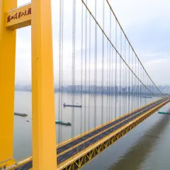 Żółty, wiszący most w Chinach przecina szeroką rzekę, po której płyną barki. W oddali widać panoramę miasta.