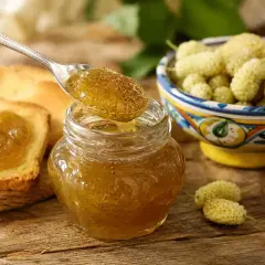 Marmellata di gelsi bianchi su pane tostato, con una ciotola di frutti freschi e un cucchiaino che ne raccoglie una porzione.