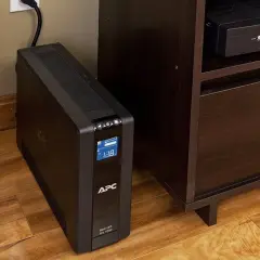 Ups co to? Czarny UPS APC Back-UPS Pro 1000 stoi na drewnianej podłodze obok mebla.