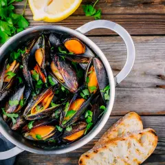 Un pentolino pieno di cozze alla marinara, guarnite con prezzemolo fresco e accompagnate da fette di pane tostato e spicchi di limone.