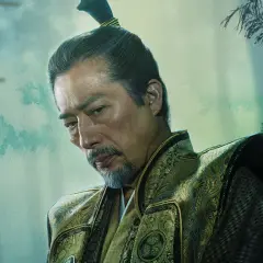 En äldre man med gråsprängt skägg och en tofs i håret, klädd i en utsmyckad dräkt. En karaktär från shogun serie.