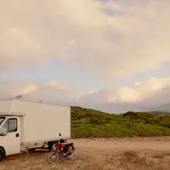 Biała kamperka i czerwony motor stoją na piaszczystej drodze. W tle zielone wzgórza i chmury. Idealne miejsce na przygodę z przewodnikiem po Korfu.