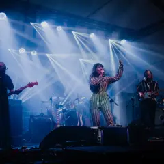 Izzy i The Black Trees na scenie, otoczeni niebieskimi światłami. Wokalistka w pasiastym stroju śpiewa, gitarzyści grają.