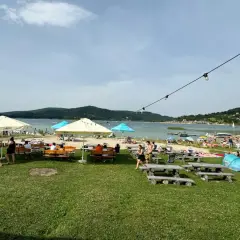 Słoneczny dzień nad Jeziorem Mucharskim. Ludzie odpoczywają na plaży, pod parasolami, przy stolikach. W tle jezioro i wzgórza. Atrakcje dla każdego.