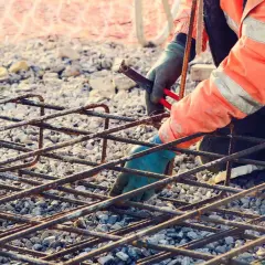 Budowlaniec w pomarańczowej kamizelce z odblaskami pracuje nad zbrojeniem rozproszonym, łącząc stalowe pręty.