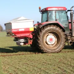 Czerwony traktor McCormick z rozsiewaczem na polu. Rozsiewacz rozprowadza saletrę wapniową, kluczową dla zdrowia roślin.