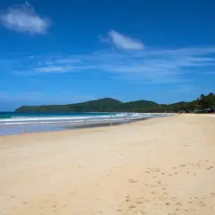 Słoneczny Nacpan Beach z turkusową wodą, zielonymi wzgórzami i palmami. Kilka osób spaceruje po piaszczystym brzegu.