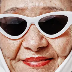 En närbild av en äldre kvinna med vit scarf och kattögon-solglasögon. Hennes hud, med sina fina linjer, liknar en **cloud skin** som fångar ljuset.