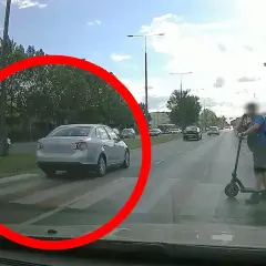 Srebrny samochód na pasach, wymuszenie pierwszeństwa. Na hulajnodze stoi mężczyzna.