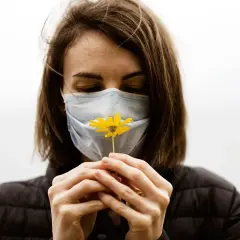 Mujer con mascarilla azul, oliendo una flor amarilla. Diversos tipos de mascarillas protegen, pero la esperanza florece.