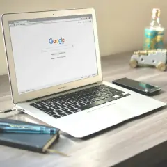 Laptop z otwartą stroną wyszukiwarki Google. Obok leży smartfon, notatnik z długopisami i ozdobne działko.