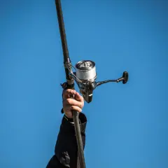 Dłoń trzyma wędkę z nawiniętą plecionką na kołowrotek, na tle błękitnego nieba.