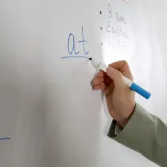 Mano scrive "at" su lavagna, spiegando il suo significato in inglese.
