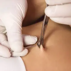 En piercare med vita handskar använder en klämma och nål för att utföra en achilles piercing.