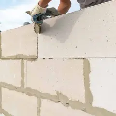 Budowa ściany z bloczków betonowych. Ręka w rękawicy nakłada zaprawę, dbając o zachowanie minimalnej grubości ściany nośnej.