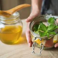 Hände halten ein Glas mit Kräutern und Zitrusfrüchten, bereit, Oxymel herzustellen. Daneben ein Glas Honig.