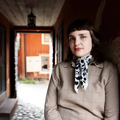 Sanna Samuelsson står i en gång med orange väggar och en träfasad i bakgrunden. Hon bär en beige tröja och en scarf med koskinnmönster.
