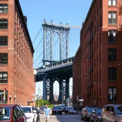 Ulica w Brooklyn, Nowy Jork, z widokiem na most Manhattan Bridge. Czerwone ceglane budynki po bokach, samochody na ulicy.