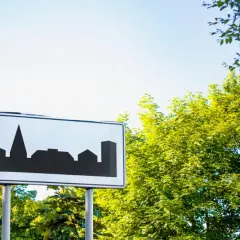 Znak D43 z sylwetką miasta. Zbliżamy się do zabudowań, a zielone drzewa szumią na tle błękitnego nieba.