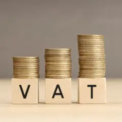 Trzy stosy monet symbolizują rosnący VAT. Litery V, A, T na kostkach tworzą napis.