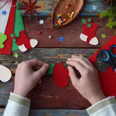 Manos creando adornos navideños con fieltro rojo y verde. Tijeras, cuentas y piñas completan la escena de manualidades.