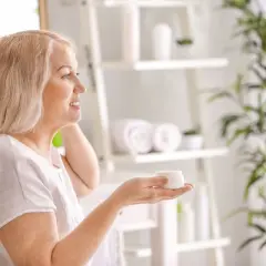 Uśmiechnięta kobieta z blond włosami trzyma słoiczek kremu, zastanawiając się, co jest dobre na cerę. W tle łazienka z roślinami.