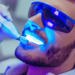 Mężczyzna w okularach ochronnych przechodzi wybielanie zębów u dentysty. Niebieskie światło lampy aktywuje żel wybielający.