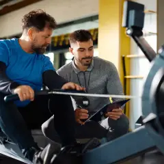 Trener crossfit omawia plan treningowy z podopiecznym na wioślarzu. Zastanawiasz się, ile zarabia trener crossfit?