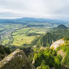 Widok z góry na dolinę z rzeką i miasteczkiem, w oddali majaczą Tatry. Widać skaliste zbocza porośnięte lasem, a w tle trzy korony nazwy szczytów.