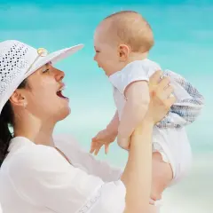 Mamma solleva il suo neonato di 5 mesi, forse le prime parole sono in arrivo, con il mare azzurro sullo sfondo.