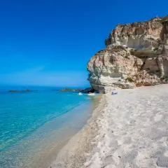 A secluded beach in Calabria, Italy, with crystal-clear turquoise water, white sand, and dramatic cliffs. A bright green umbrella offers shade on this idyllic spot.