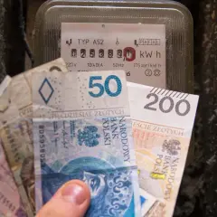 Dłoń trzyma polskie banknoty przy liczniku energii elektrycznej. Zastanawiasz się, po ile jest prąd?